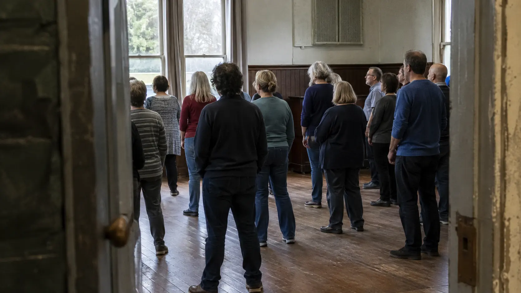Adults stand in a loose group facing one side of an older multipurpose room.