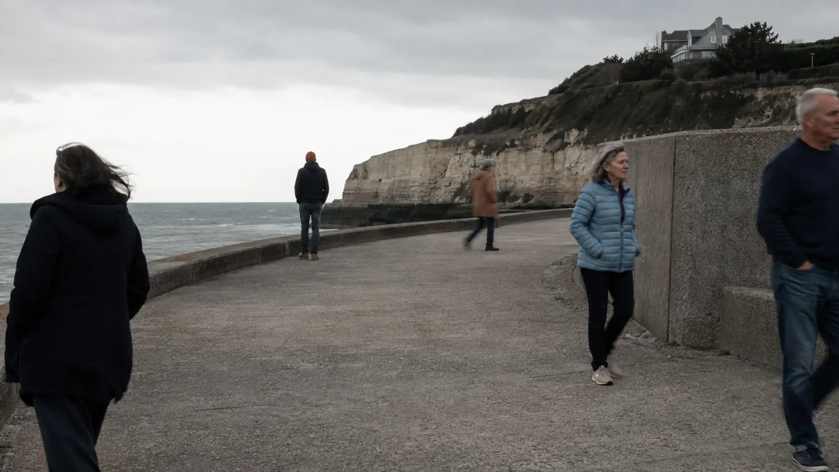 Several adults walk in different directions on a weathered seafront promenade under cloud.