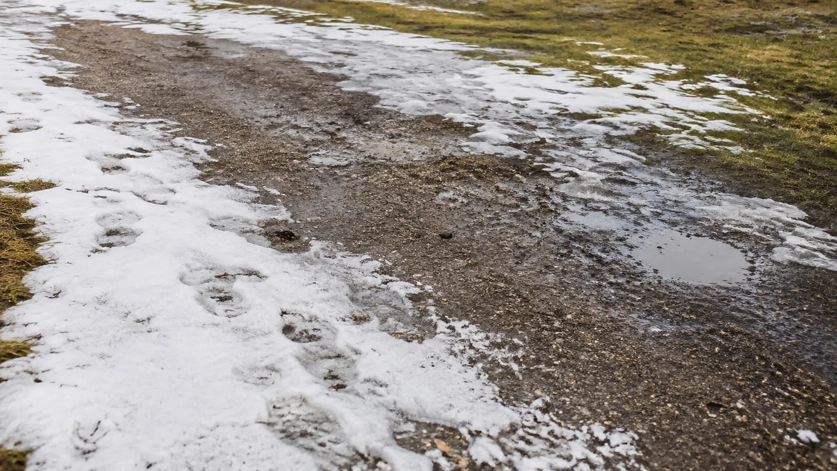 Partly softened footprints overlap across thawing snow, gravel and grass.